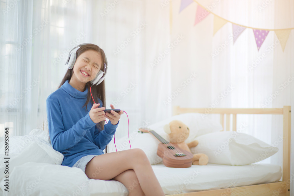 Pretty Asian girl using headphone for listen music by smartphone on the bed in her decorated bedroom