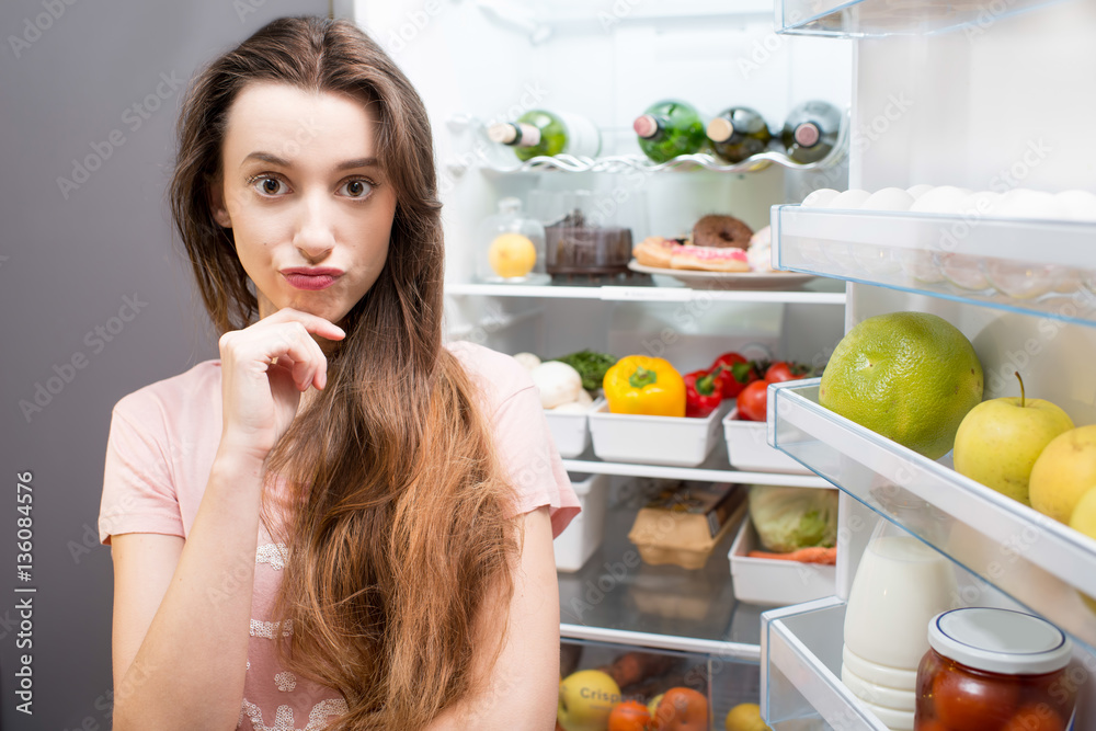 Portrait of a young thinking woman in front of the refrigerator full of food