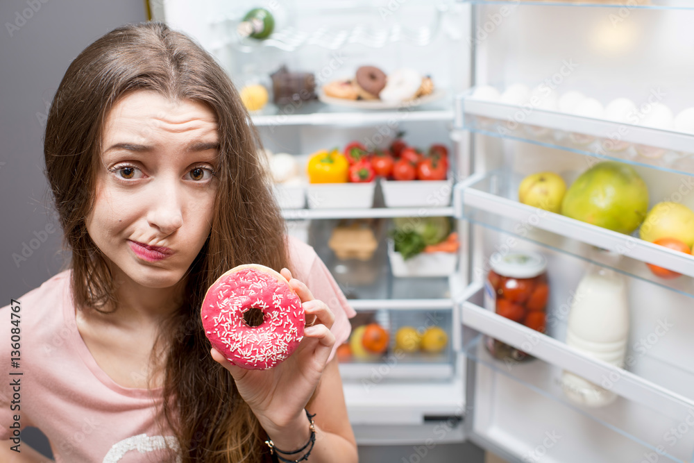 Portrait of a young sad woman in the sleepwear with sweet donuts standing near the open refrigerator