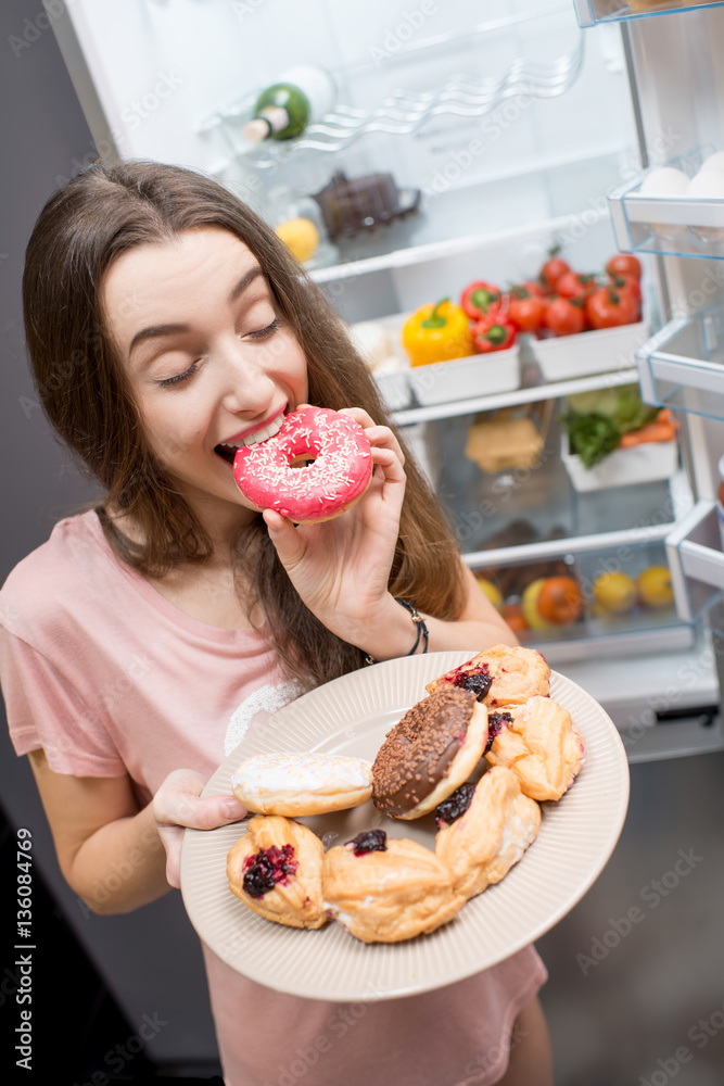 Young woman in the sleepwear eating sweet donuts near the refrigerator