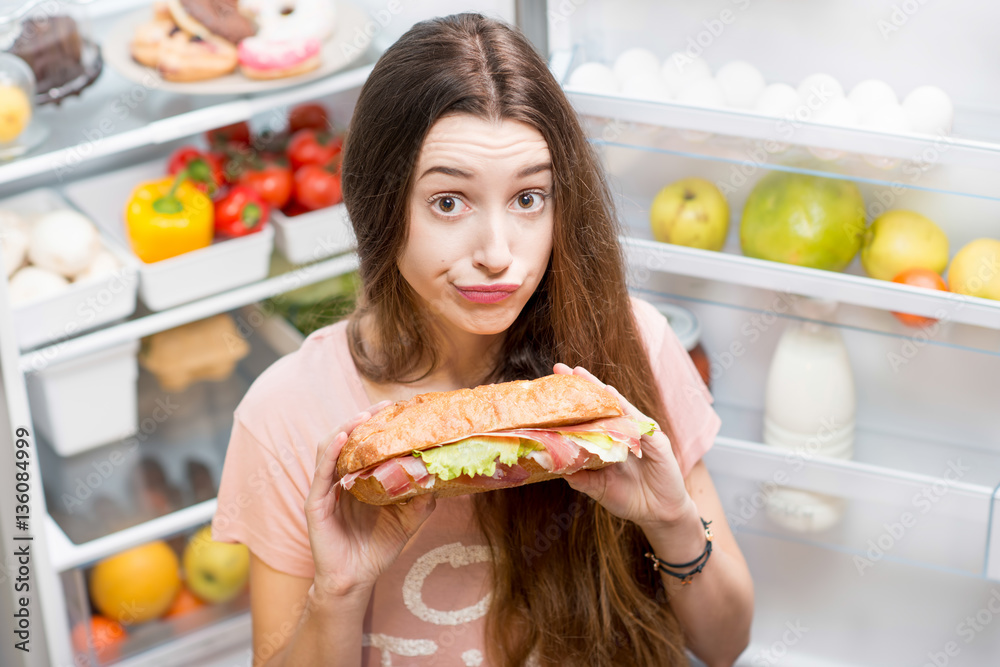 Young woman with big sandwich in front of the refrigerator full of friuts and vegetables