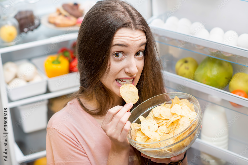 Young woman eating potato chips near the refrigerator full of healthy food