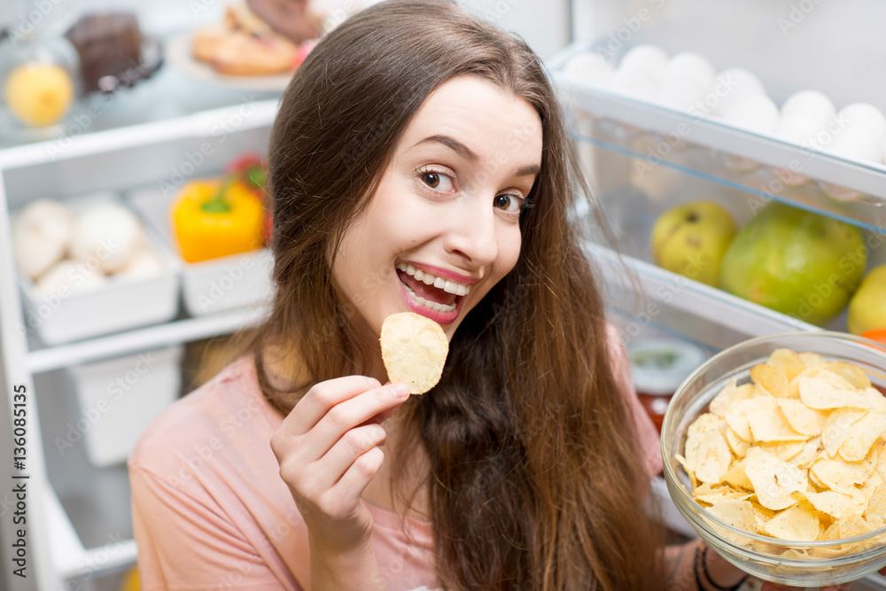 Young woman eating potato chips near the refrigerator full of healthy food