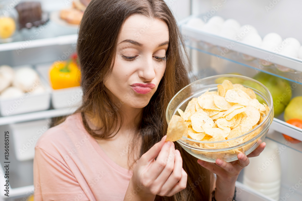 Young woman eating potato chips near the refrigerator full of healthy food