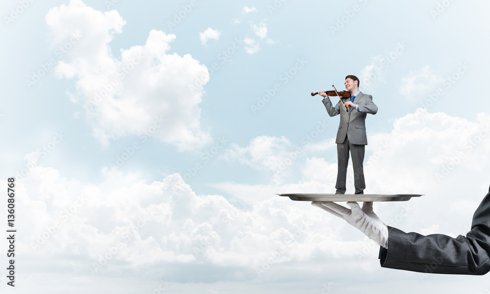 Businessman on metal tray playing violin against blue sky background