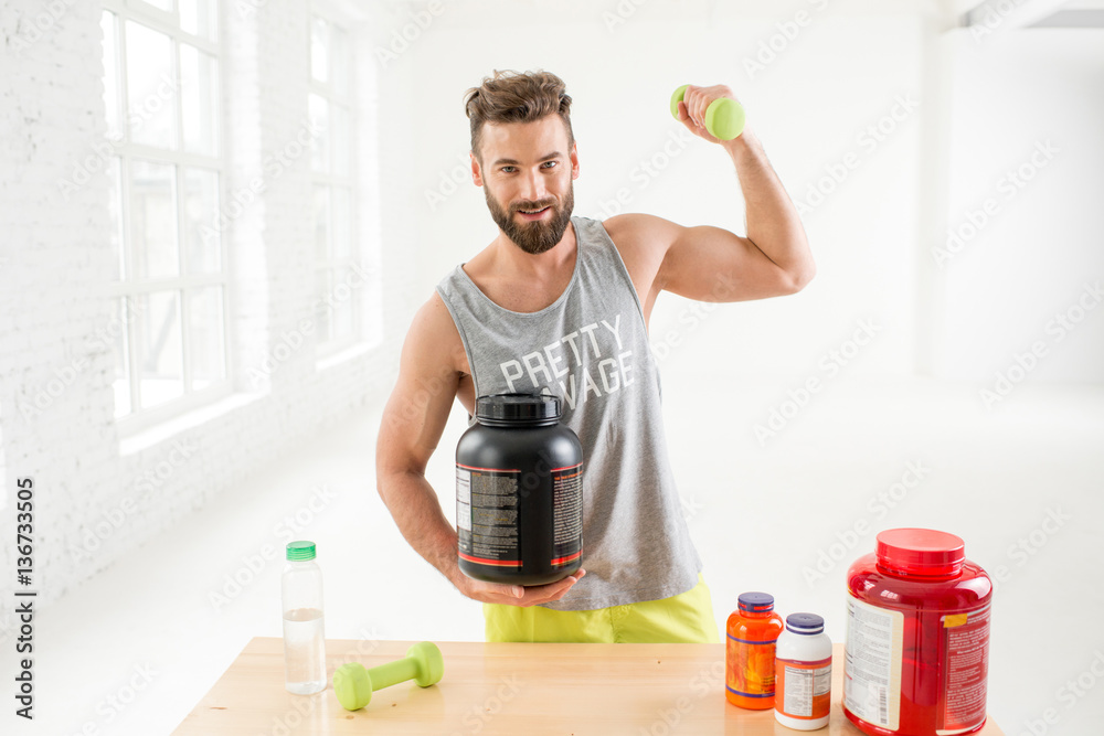 Athletic man training with dumbbells with proteins and vitamins on the table in the white gym interi