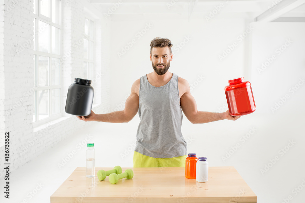 Athletic man holding bottles with sports nutriton in the white gym interior. Blank bottles to copy p