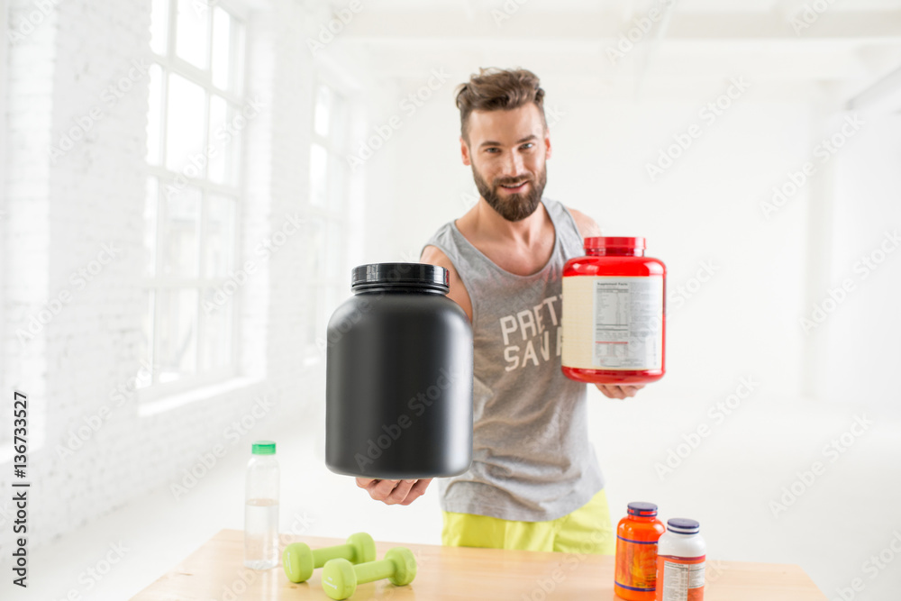 Athletic man holding bottles with sports nutriton in the white gym interior. Blank bottles to copy p