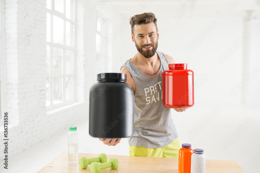 Athletic man holding bottles with sports nutriton in the white gym interior. Blank bottles to copy p