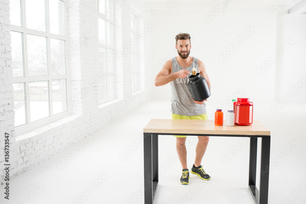 Athletic man in sportswear pouring with scooper protein powder in the white gym interior