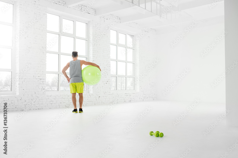 Wide shot of the white gym interior with big windows and man standing with fitball