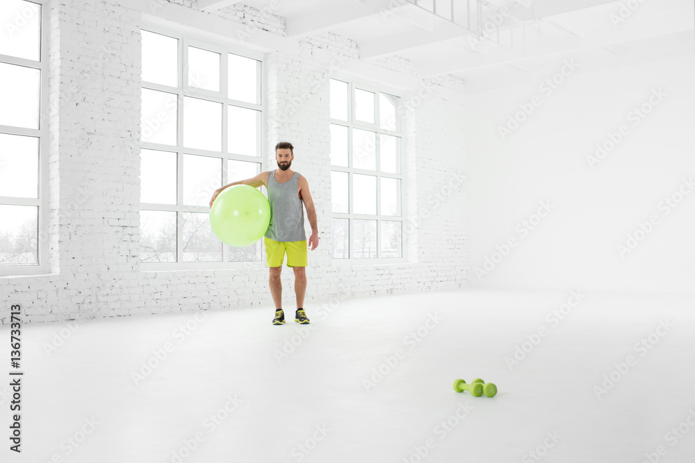 Wide shot of the white gym interior with big windows and man standing with fitball