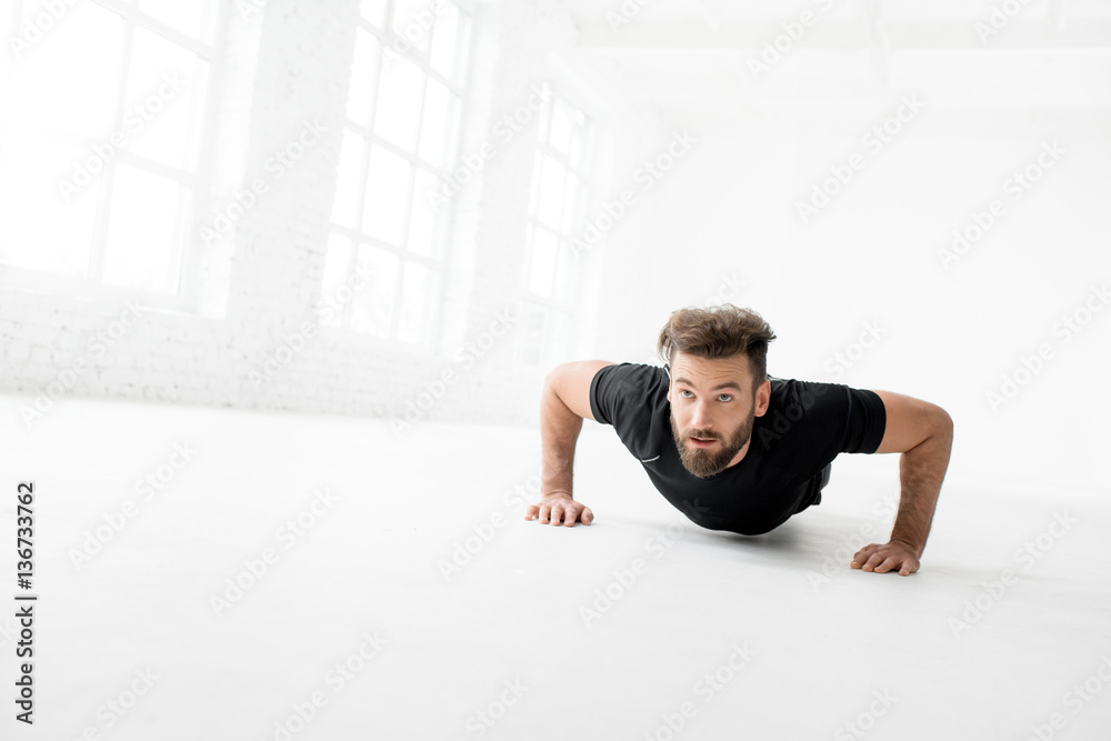 Handsome man in the black sportswear making pushups indoors in the white gym interior