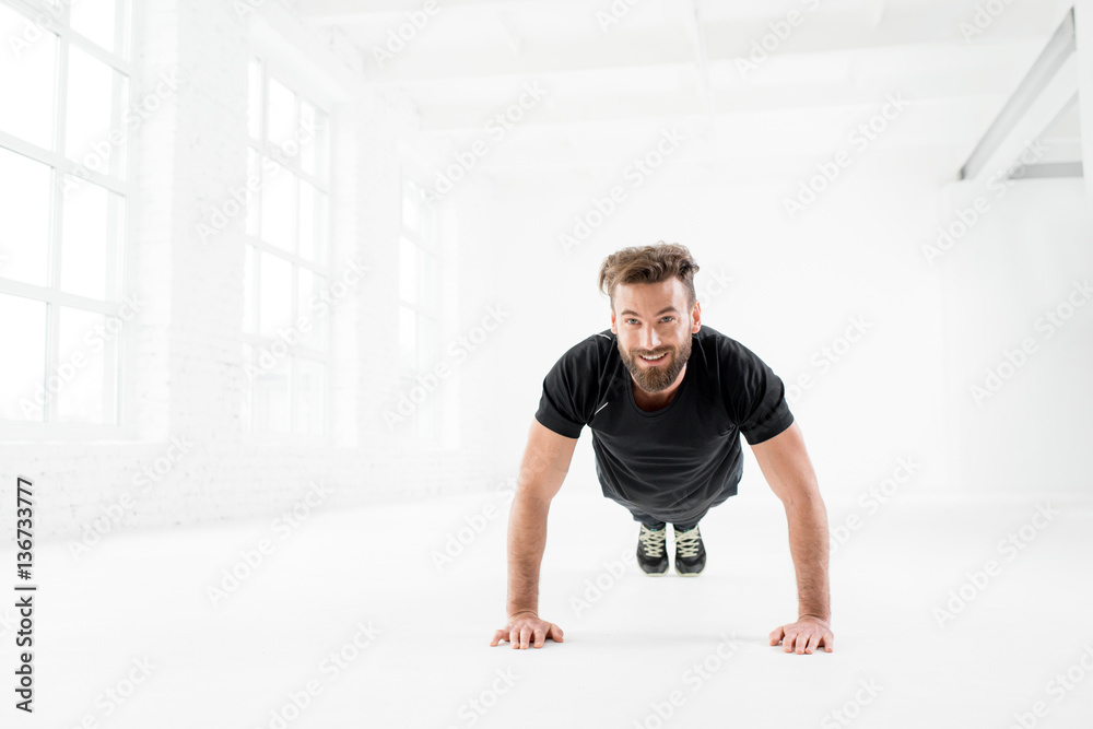 Handsome man in the black sportswear making pushups indoors in the white gym interior