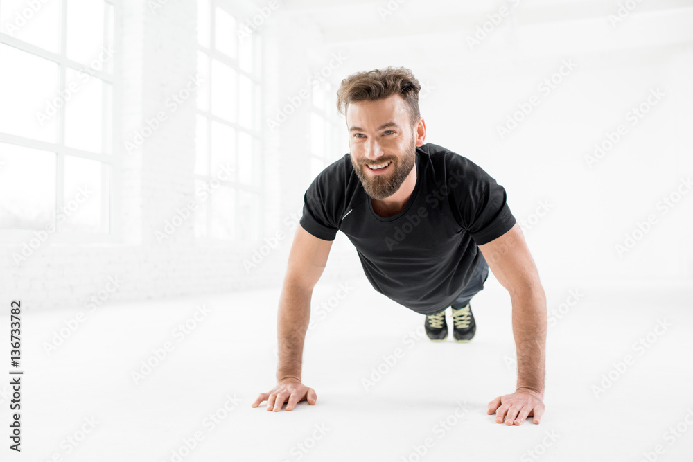 Handsome man in the black sportswear making pushups indoors in the white gym interior