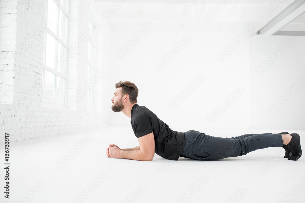 Handsome man in the black sportswear doing yoga in the white gym interior