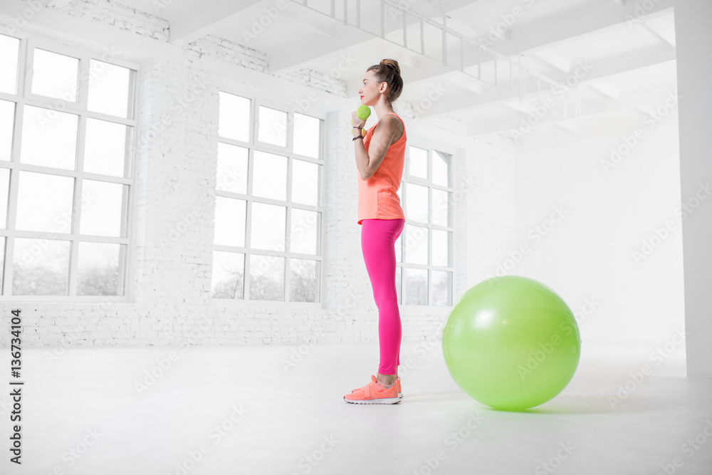 Young woman in colorful sportswear lifting dumbbels in the white gym with green fitness ball on the 