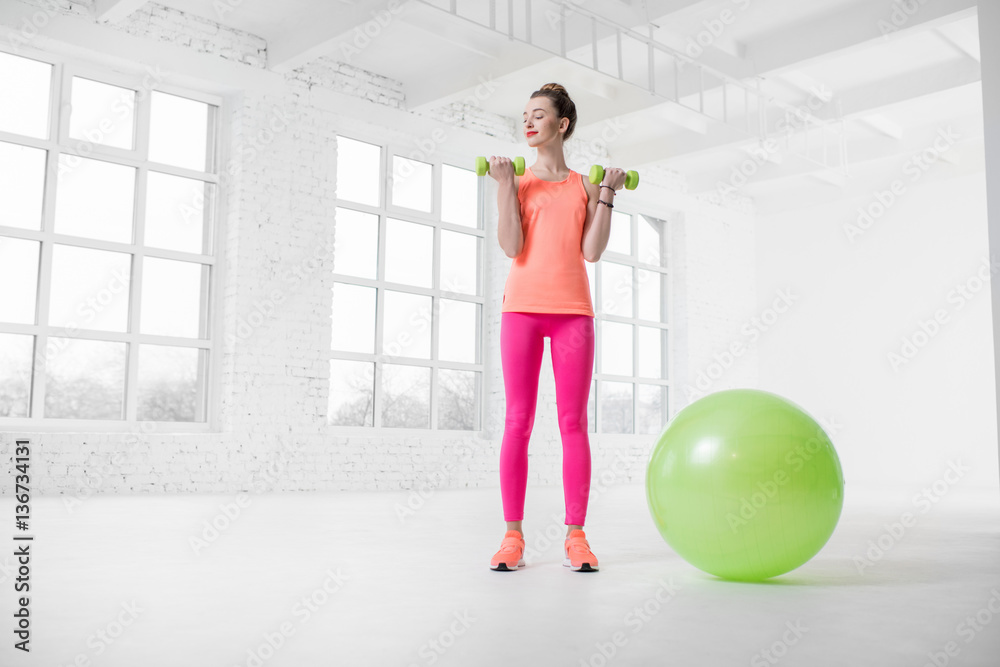 Young woman in colorful sportswear lifting dumbbels in the white gym with green fitness ball on the 