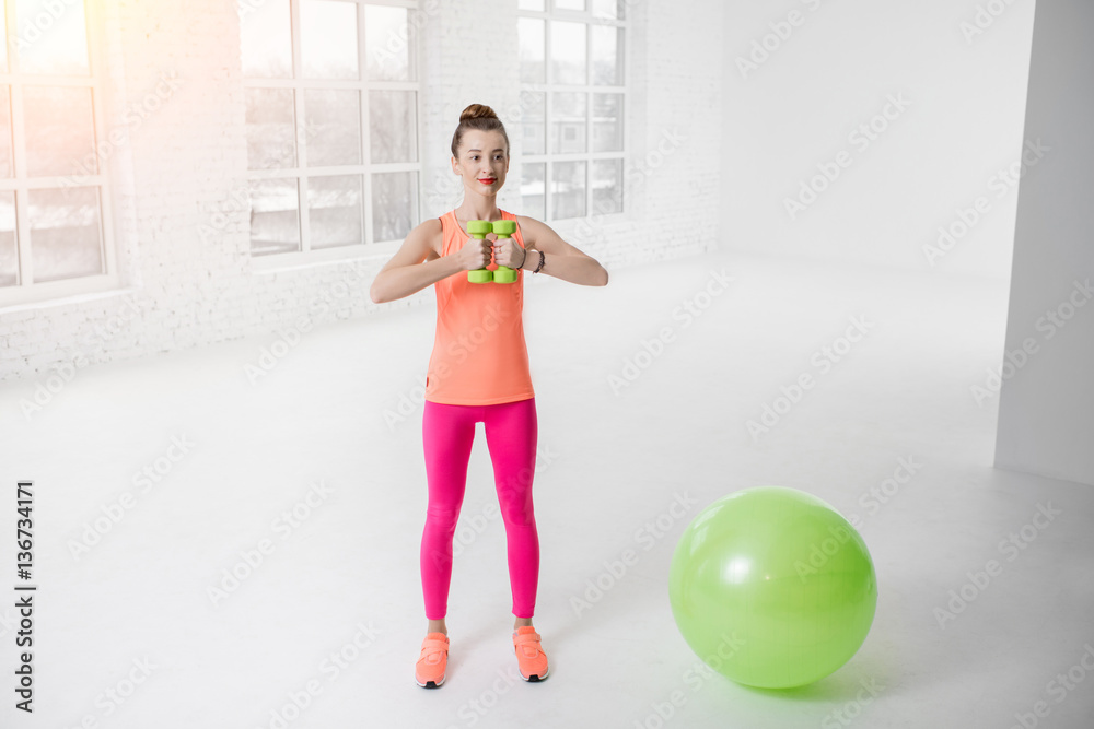 Young woman in colorful sportswear lifting dumbbels in the white gym with green fitness ball on the 