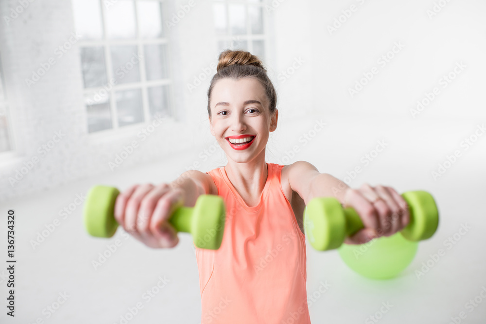 Portrait of young woman in sportswear with dumbbells and fitness ball in the white gym