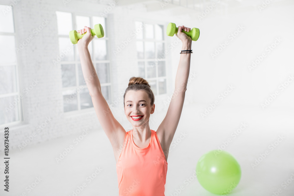 Portrait of young woman in sportswear with dumbbells and fitness ball in the white gym