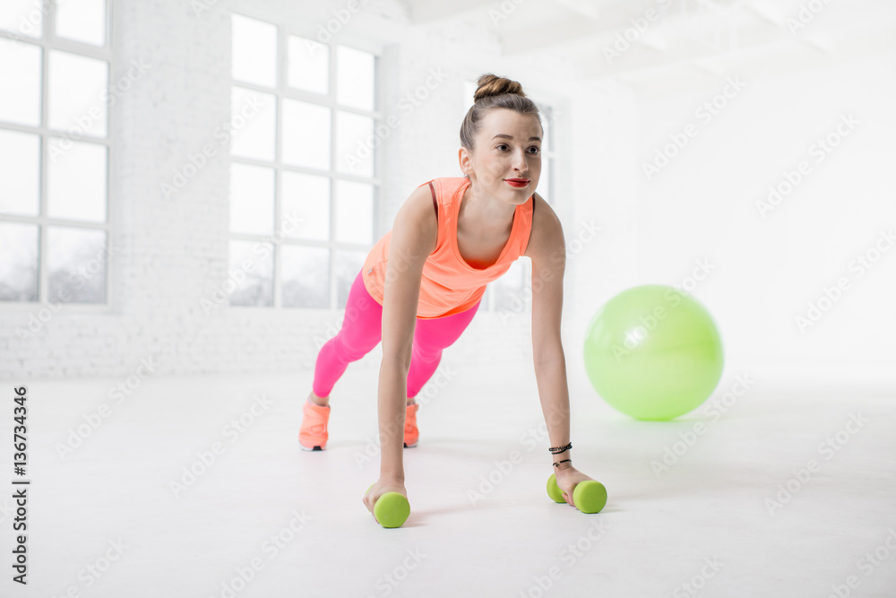 Young woman in colorful sportswear making push ups with dumbbels and fitness ball on the background