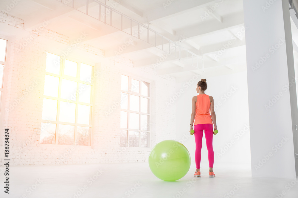 Young woman in colorful sportswear standing with dumbbels in the white room with green fitness ball 