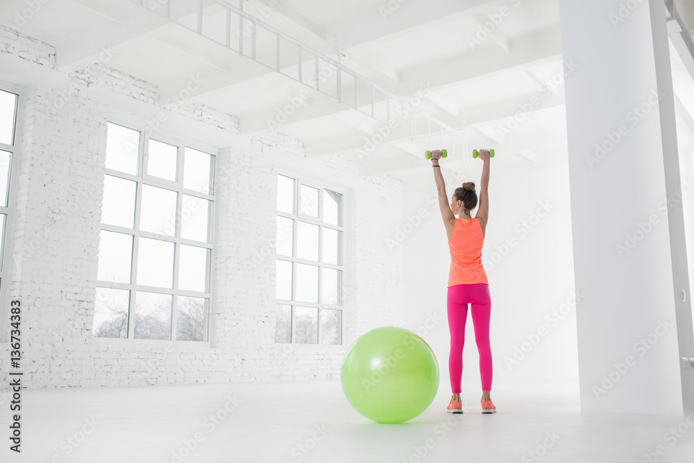 Young woman in colorful sportswear lifting dumbbels in the white room with green fitness ball on the