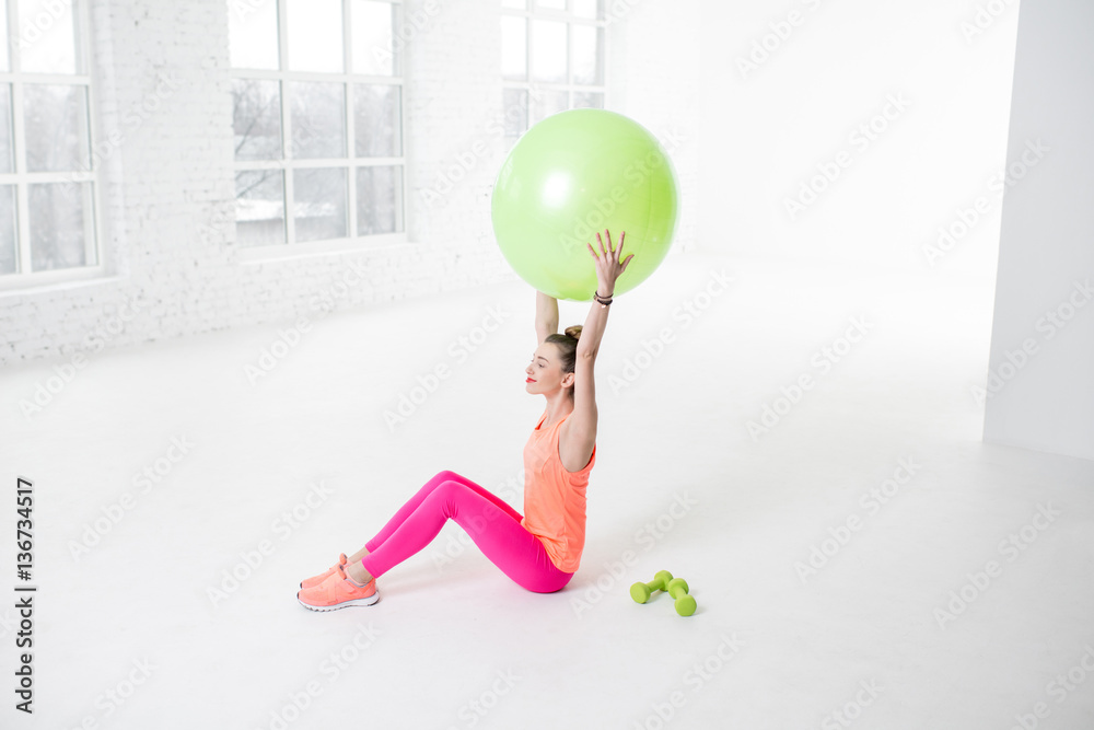 Young woman in colorful sportswear stretching with fitness ball in the white gym