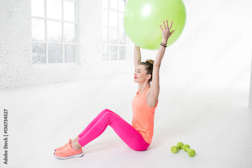 Young woman in colorful sportswear stretching with fitness ball in the white gym