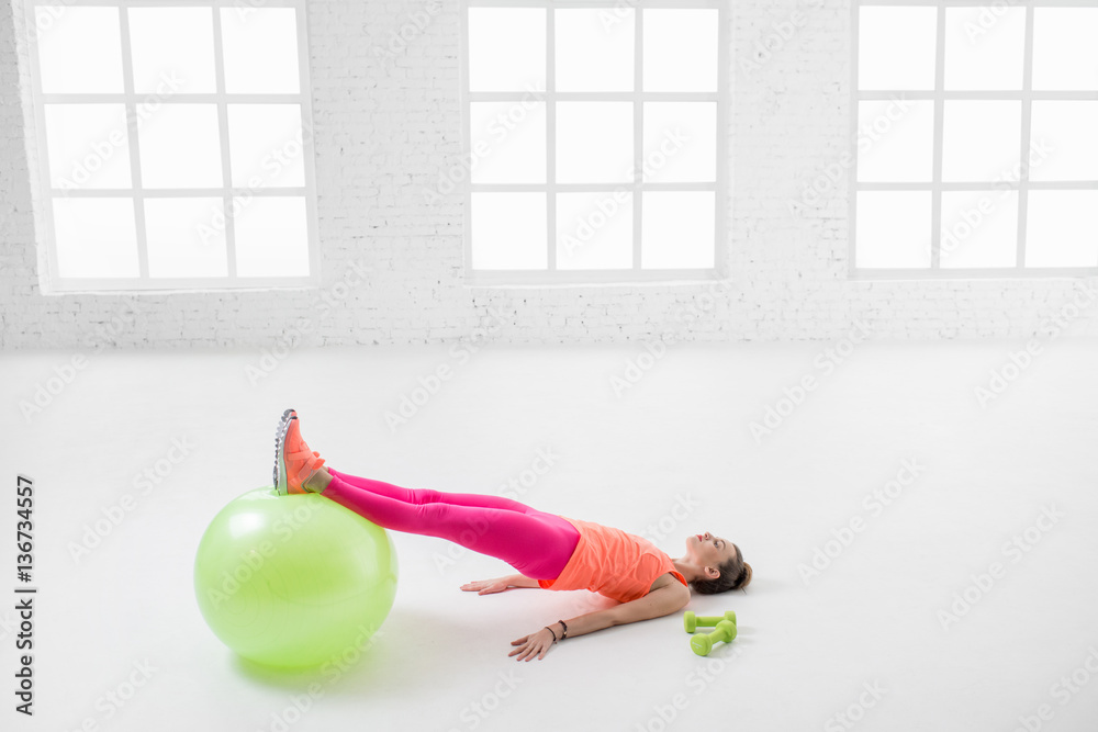 Young woman in colorful sportswear stretching with fitness ball in the white gym