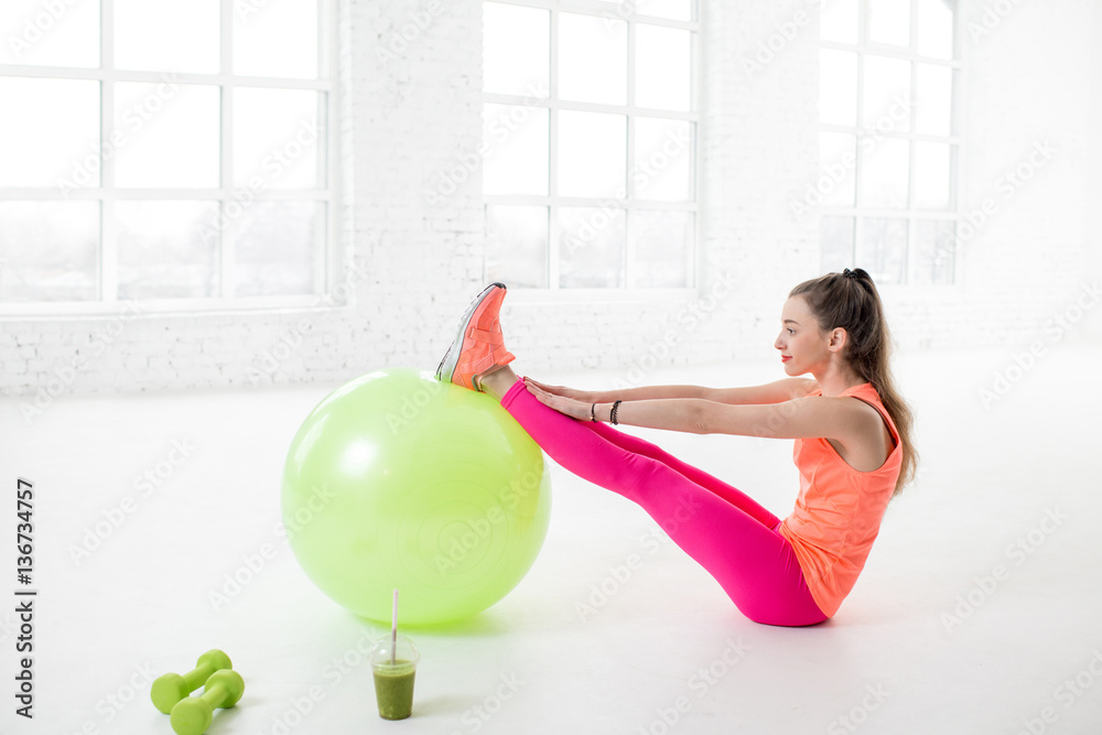 Young woman in colorful sportswear stretching with fitness ball in the white gym