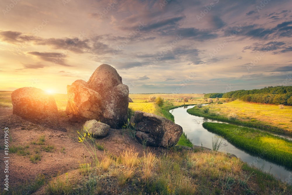 Summer landscape at sunset. Big stones at the river against blue sky with clouds and yellow sunlight