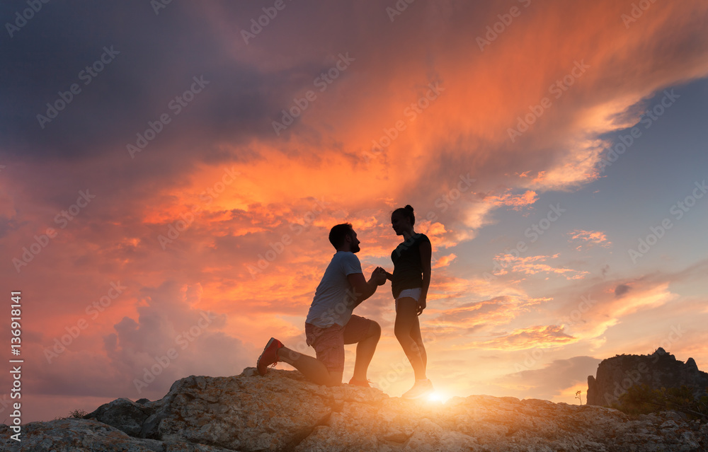 Silhouettes of a man making marriage proposal to his girlfriend on the mountain peak at sunset. Land