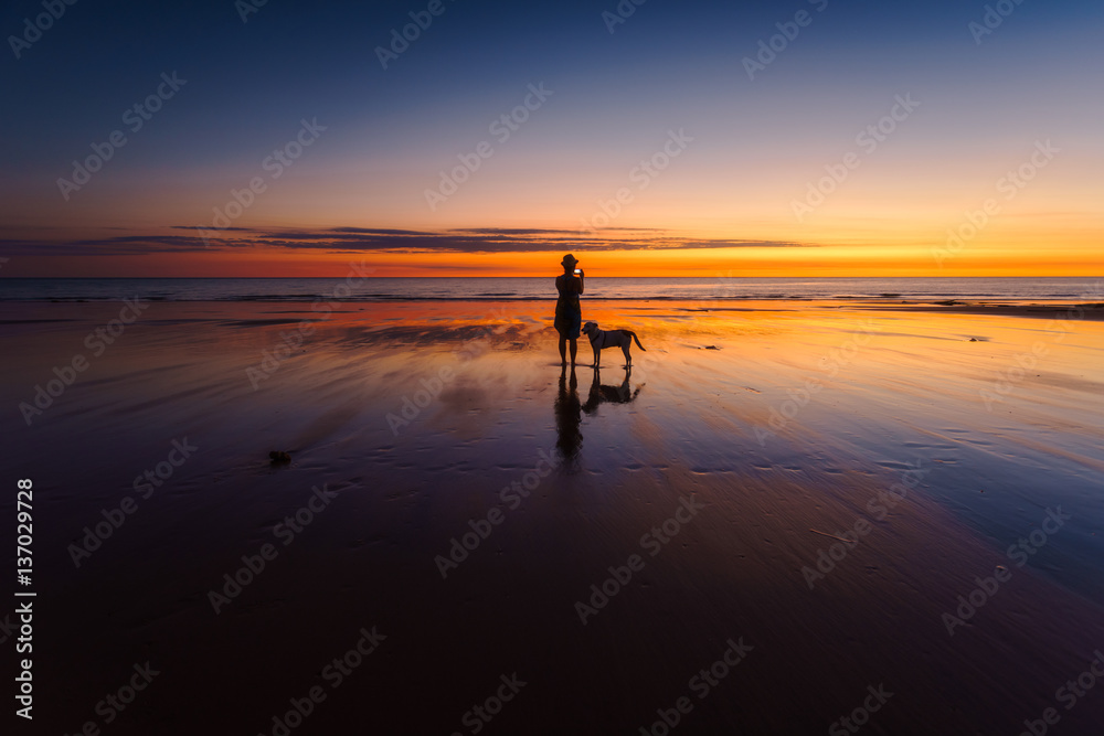 A woman taking a photograph of the sunset is silhouetted with her dog beside her. Western Australia,