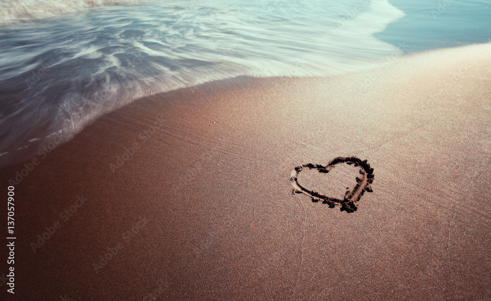 Lovely heart symbol hand drawn on the sunny sandy beach with wave. Selective focus used.