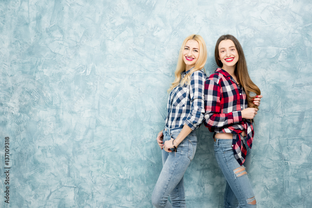 Portrait of two female friends in checkered shirts and jeans together on the blue painted wall backg