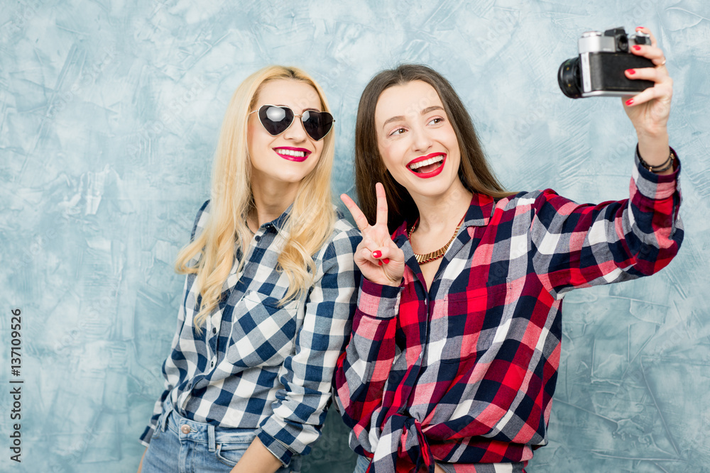 Two female friends in checkered shirts and jeans photographing with retro camera on the blue painted