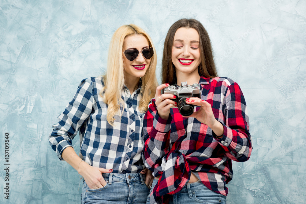 Two female friends in checkered shirts and jeans photographing with retro camera on the blue painted