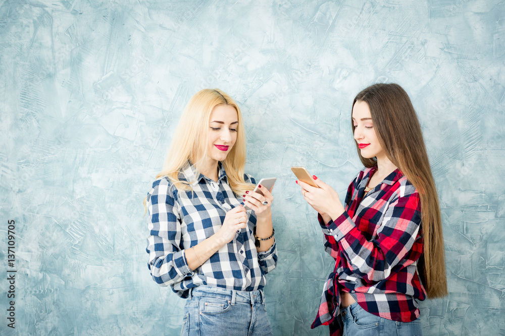 Female friends in checkered shirts using smart phones on the blue wall background