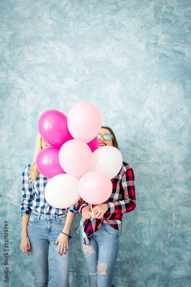 Two female friends in checkered shirts having fun with air balloons on the blue wall background