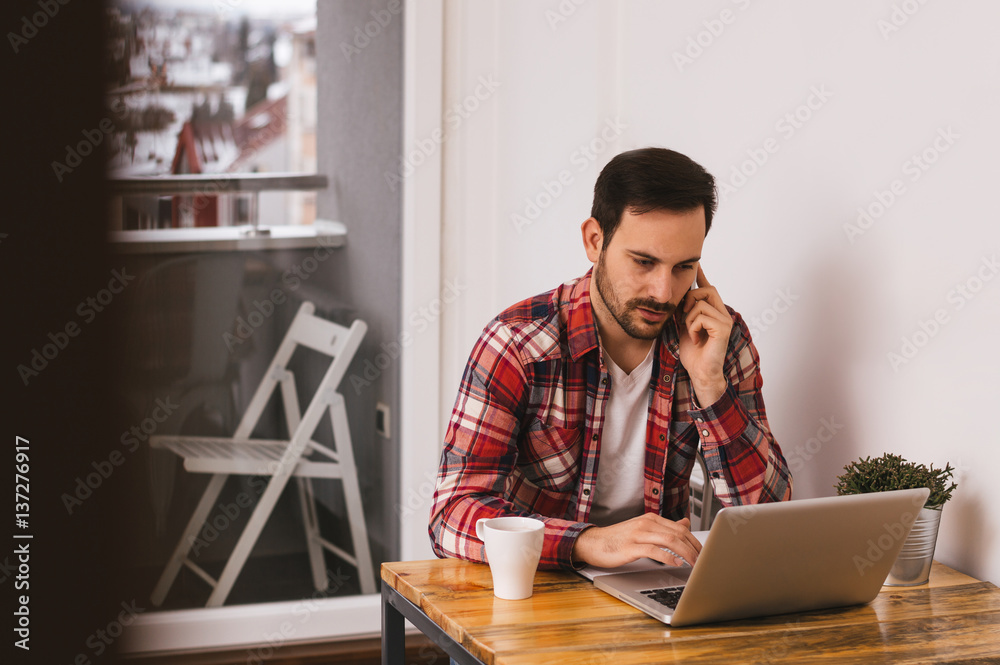 Handsome man working on laptop at home office, talking on the phone.