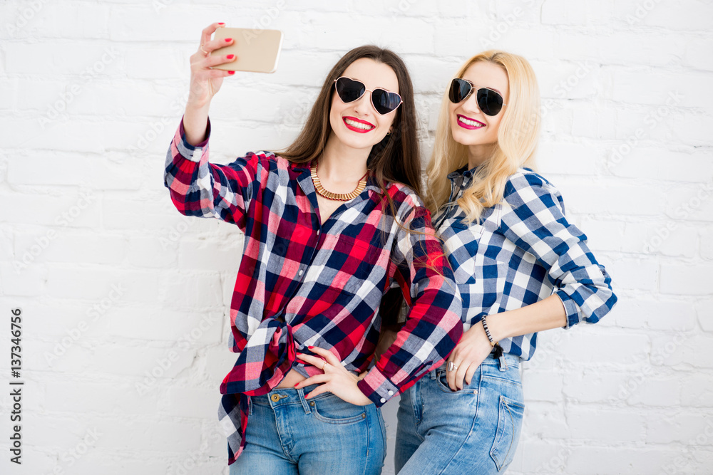 Female friends in checkered shirts making selfie portrait with phone on the white wall background