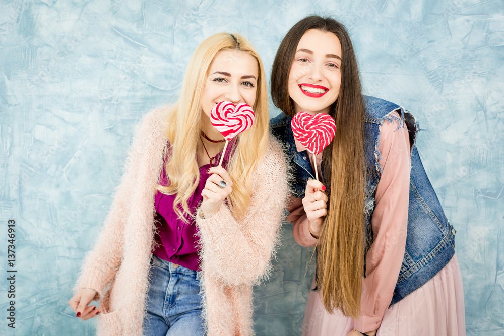 Female stylish friends having fun with candy on the blue wall background