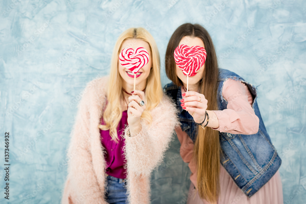 Female stylish friends having fun with candy on the blue wall background