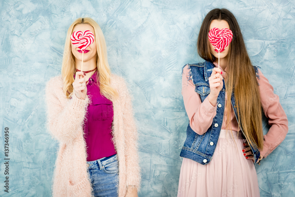 Female stylish friends having fun with candy on the blue wall background