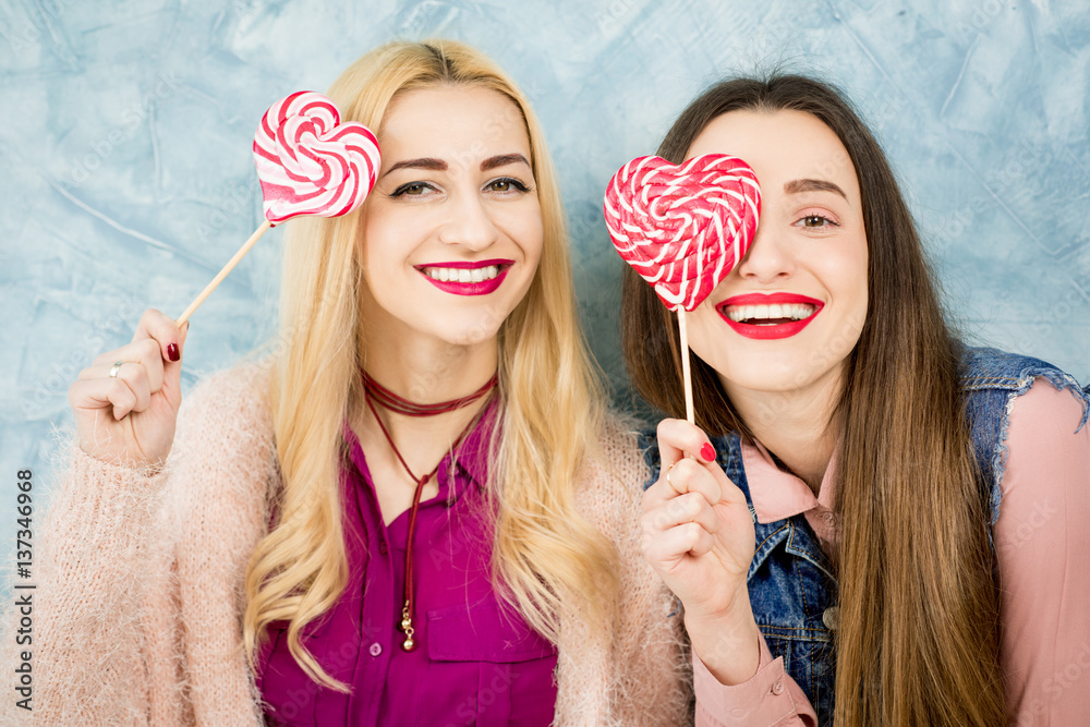 Female stylish friends having fun with candy on the blue wall background