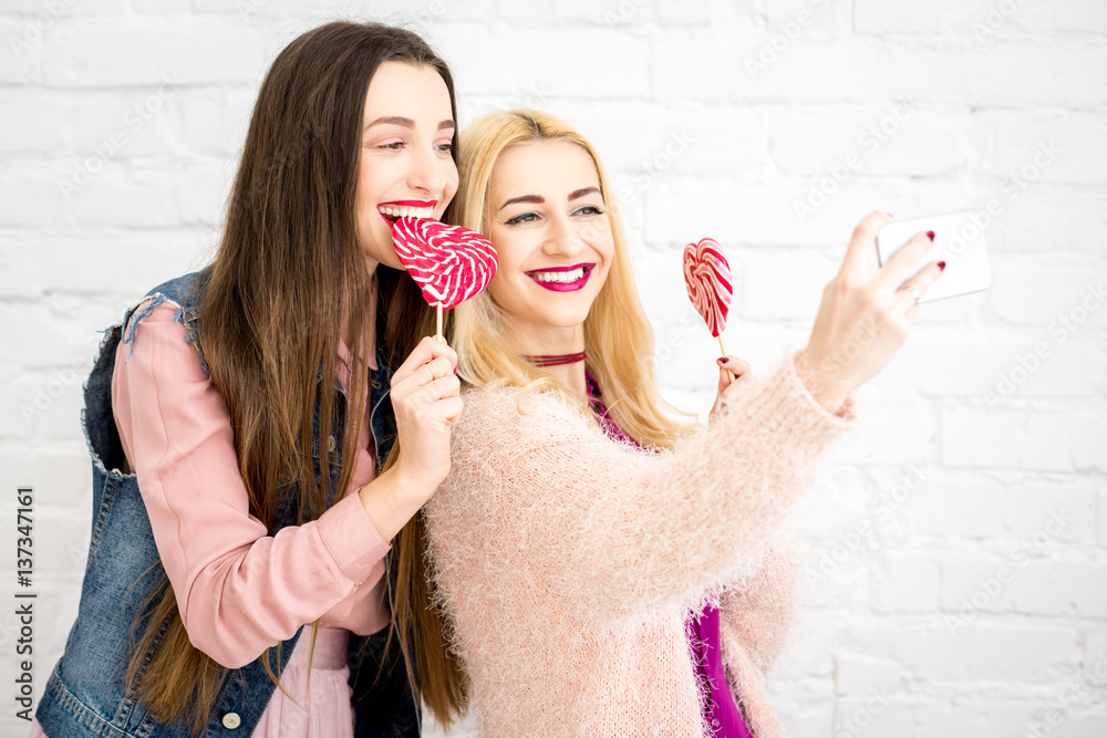 Female stylish friends making selfie photo with red candies on the white wall background