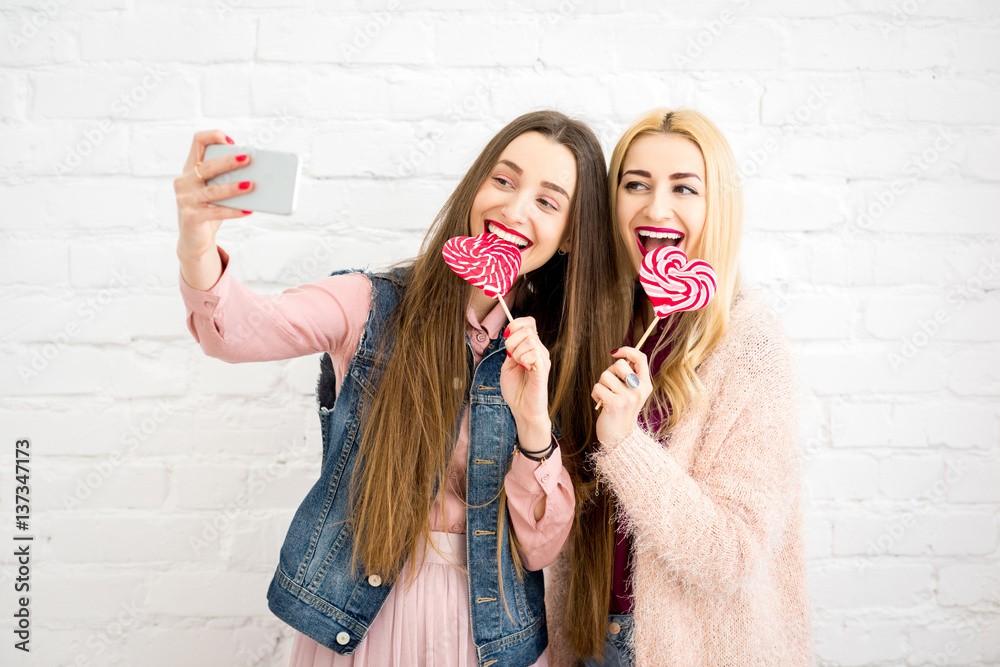 Female stylish friends making selfie photo with red candies on the white wall background