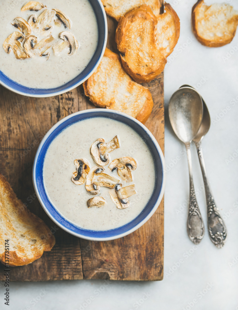 Close-up of creamy mushroom soup in bowls with toasted bread slices on rustic serving board over gre